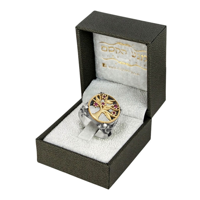 Puede incluir: Un anillo de plata con un dise&ntilde;o de &aacute;rbol de la vida en oro y piedras preciosas rojas. El anillo est&aacute; en una caja de terciopelo verde con las palabras "Tree of Life Jewelry" en el interior.