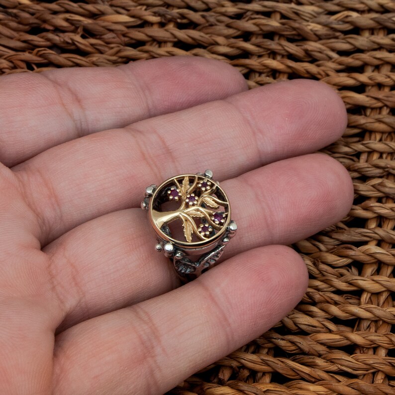 Puede incluir: Un anillo de plata con un dise&ntilde;o de &aacute;rbol de oro y piedras preciosas rojas. El &aacute;rbol est&aacute; rodeado por un c&iacute;rculo de plata con un borde texturizado.