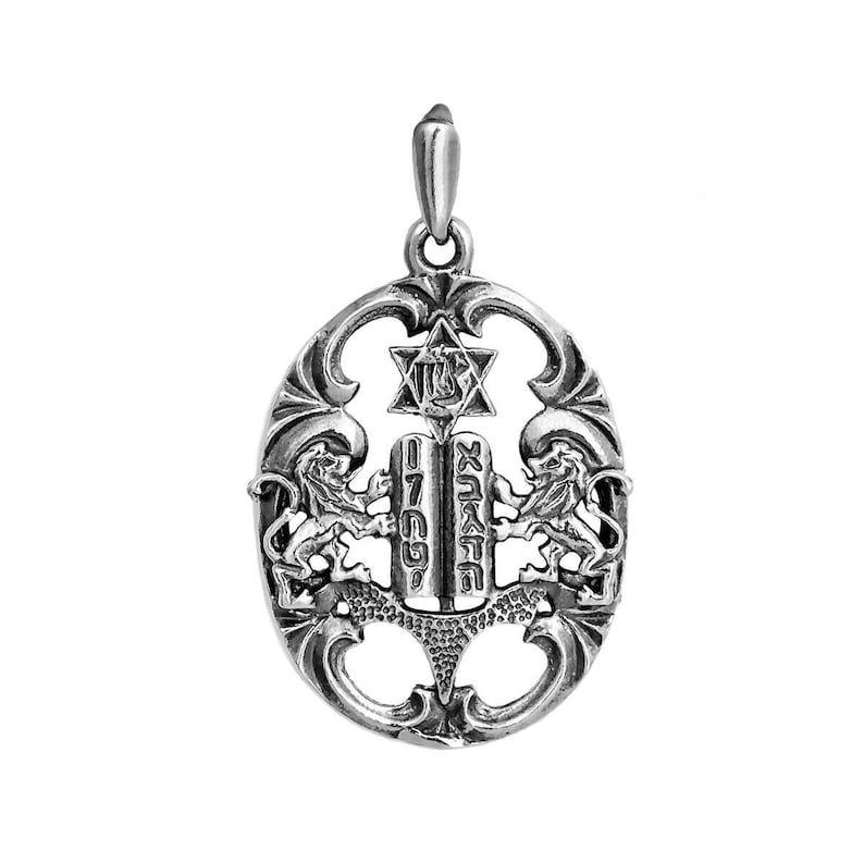 May include: An oval-shaped silver pendant featuring an openwork design with two lions flanking a central tablet inscribed with Hebrew characters. Above the tablets is a Star of David with a central symbol. The piece includes a top bail for hanging.