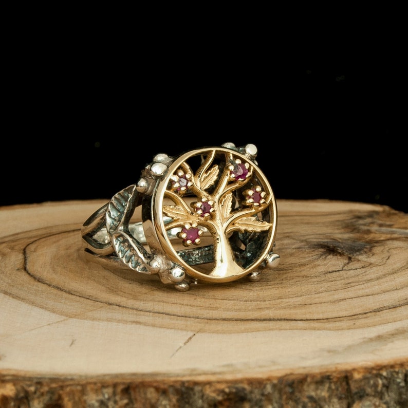 Puede incluir: Un anillo de plata con un dise&ntilde;o de &aacute;rbol de la vida en oro y piedras de rub&iacute; rojas. El anillo est&aacute; colocado sobre una superficie de madera.