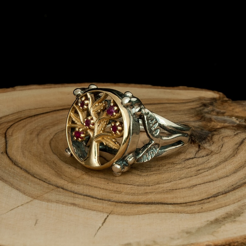 Puede incluir: Un anillo de plata con un dise&ntilde;o de &aacute;rbol de la vida en oro y piedras preciosas rojas. El anillo est&aacute; sobre una superficie de madera.