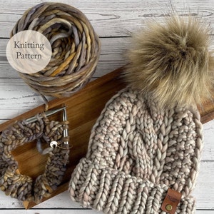 Op de afbeelding: Een beige gebreide beanie met een grote bruine nep bont pompon. De beanie heeft een kabelbreipatroon en een bruin lederen label met de tekst "The Walnut Weaver". De beanie ligt op een houten oppervlak met een bol wol en breinaalden.