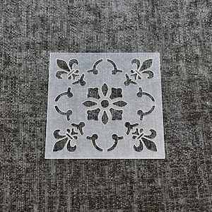 May include: A clear plastic stencil with a square design featuring a repeating pattern of stylised flowers and fleur-de-lis.