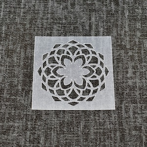 May include: A white plastic stencil featuring a black mandala design. The mandala is intricate and detailed, with a circular pattern of overlapping shapes.