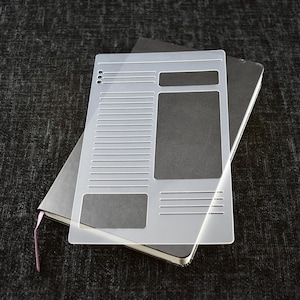 May include: A clear plastic planner stencil with a grid layout, a large rectangle, and a smaller rectangle. The stencil is placed on top of a black notebook.
