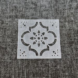 May include: A white plastic stencil with a geometric design featuring a central flower motif. The stencil is ideal for creating decorative patterns on walls, furniture, or crafts.