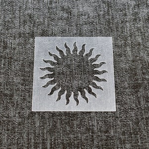 May include: A clear plastic stencil featuring a sun design. The sun has a circular shape with rays extending outwards.
