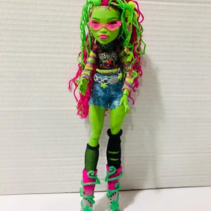 Peut inclure: Poupée à la peau verte avec des cheveux roses et verts, portant des lunettes de soleil roses, un t-shirt noir avec le texte "NATURE", un short en jean et des bottes à plateforme argentées. La poupée a des jambières noires et vertes et des accents roses et verts en forme de vigne.