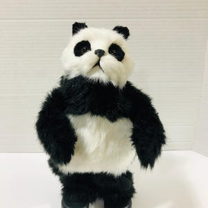 Może przedstawiać: Pluszowa zabawka panda z białą twarzą, czarnymi plamami wokół oczu, czarnym nosem i białą klatką piersiową. Ciało pokryte jest czarno-białym sztucznym futrem, a zabawka stoi na dwóch okrągłych, czarnych plastikowych stopach u podstawy.