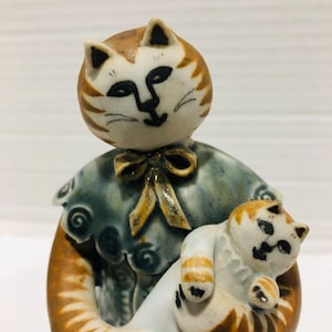May include: A ceramic figurine of a cat holding a smaller cat. The larger cat is brown and white with a blue and white patterned dress. The smaller cat is white with brown stripes.