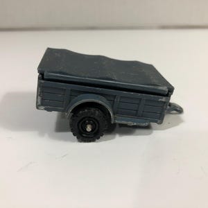 Puede incluir: Un pequeño remolque de juguete gris con un neumático negro y una cubierta gris oscuro. El remolque tiene forma rectangular con paneles laterales de estilo madera y un pequeño enganche. El juguete está diseñado para ser tirado por un vehículo.