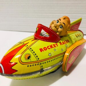 May include: A vintage tin toy rocket racer in yellow, red, and orange. The words "ROCKET RACER" are in red. A smiling figure is in the cockpit. The rocket has a retro design with stars and a planet on the side.