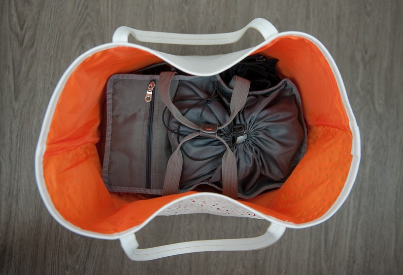 May include: A white tote bag with orange lining and a gray drawstring bag inside. The tote bag has a white handle and a white zipper.