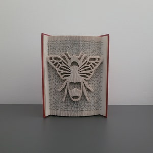 Könnte beinhalten: Ein Buch mit einem bienenförmigen Ausschnitt, der durch das Falten der Seiten entstanden ist. Das Buch ist rot und die Biene ist weiß.