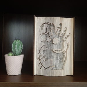 May include: A book art sculpture of a dragon made from the pages of a book. The sculpture is displayed on a wooden shelf with a small potted cactus in front of it.