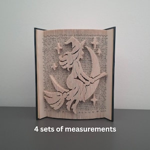 May include: A carved book sculpture featuring a witch riding a broom across a crescent moon, with stars. The artwork is made from the pages of a book, with the edges of the pages forming the image. The book is open and standing upright.
