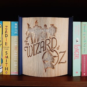 May include: A bookend featuring a carved design of "The Wizard of Oz" characters, made from a book with pages folded into a 3D design. The bookend is displayed on a bookshelf with other books, including "White Teeth" by Zadie Smith, "Swing Time" by Zadie Smith, "Becoming Jo" by Meg Keene, "This is Going to Hurt" by Adam Kay, and "The Choice" by Samantha King.