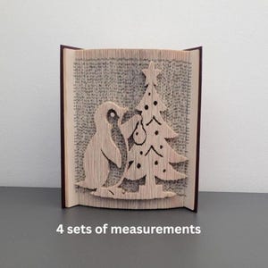 May include: A book sculpture with a penguin and Christmas tree design, created by folding the pages. The book has a dark brown cover. The text "4 sets of measurements" is visible at the bottom of the image.