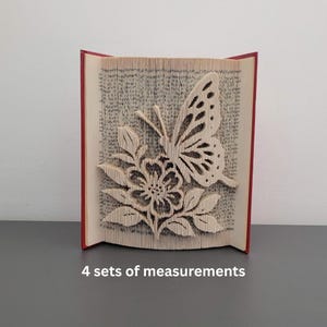 Peut inclure: Sculpture de livre avec un papillon et une fleur. Les pages sont coupées et pliées pour créer un effet 3D. Le livre a une tranche rouge et le texte "4 sets of measurements" est en bas.