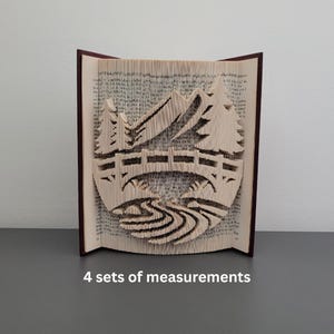 May include: A book sculpture depicting a mountain scene with a bridge and trees. The pages are intricately cut to form the design, with a dark burgundy cover. The book is displayed upright on a grey surface.