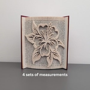 Könnte beinhalten: Ein Buchskulptur mit einem geschnitzten Hibiskusblüten-Design. Die Seiten sind gefaltet, um die Blume zu bilden, mit einem weinroten Einband. Der Text "4 sets of measurements" ist unten im Bild zu sehen.