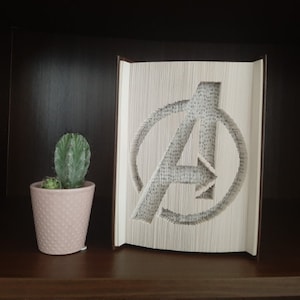 以下が含まれることがあります： アベンジャーズのロゴの形に折り畳まれた白い本。円の中に「A」の文字。本は濃い茶色の棚の上にあり、その前に小さな鉢植えの サボテン が置かれている。