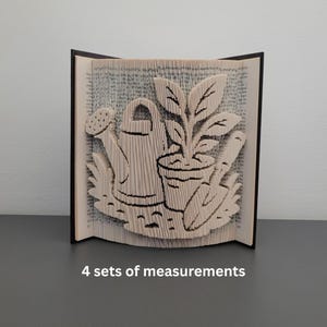 May include: A book sculpture featuring a garden scene with a watering can, potted plant, and gardening tools. The text "4 sets of measurements" is visible at the bottom. The sculpture is made from the pages of a book.