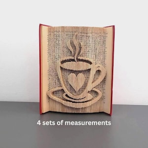 May include: A book sculpture with a coffee cup design. The pages are folded to create a 3D image of a coffee cup with a heart in the center. The book has a red cover and is displayed on a gray surface.