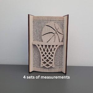 Puede incluir: Escultura de arte de libro única con un diseño de baloncesto y aro. El balón de baloncesto está colocado encima del aro, ambos meticulosamente elaborados a partir de las páginas de un libro. El libro tiene una cubierta marrón oscuro y se muestra en posición vertical.