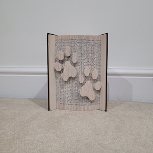 Op de afbeelding: Een boek met pagina's gevouwen in de vorm van twee poot afdrukken. Het boek is gesloten en staat rechtop op een lichtbruin tapijt.