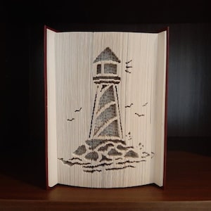 May include: A white book with pages folded into a 3D design of a lighthouse on a rocky shore. The book is standing upright on a wooden shelf.