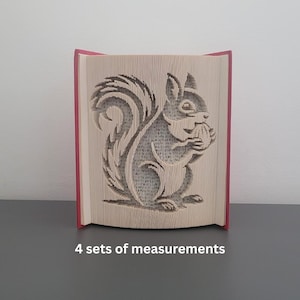 Może przedstawiać: Rzeźba z książki z wyrzeźbionym wzorem wiewiórki trzymającej orzech. Wiewiórka jest wycięta ze stron książki, z widocznymi krawędziami stron. Książka ma czerwone okładki i stoi pionowo.