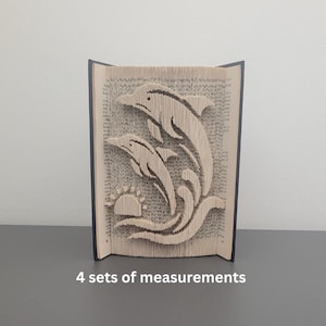 Op de afbeelding: Een boeksculptuur met twee dolfijnen die over golven en een zon springen. Het ontwerp is uit de pagina's van een boek gesneden, waardoor een 3D-effect ontstaat. Het boek is open, met het gesneden beeld zichtbaar. De tekst "4 sets of measurements" staat onderaan.