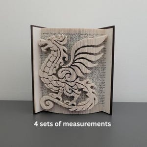 Könnte beinhalten: Buchskulptur mit einem detaillierten Drachendesign, das in die Seiten geschnitten wurde. Der weiße Drache hat detaillierte Flügel und einen Körper. Das Buch ist geöffnet und zeigt den Drachen. Der Text "4 sets of measurements" befindet sich unten.