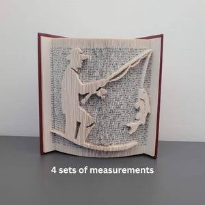 Op de afbeelding: Een unieke boekkunstsculptuur die een visser afbeeldt die een vis vangt. Het kunstwerk is gemaakt door de pagina's van een boek ingewikkeld te vouwen. Het boek heeft een rode rug en de tekst "4 sets of measurements" staat onderaan.
