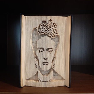 May include: A book sculpture featuring a portrait of a woman with a floral crown and earrings. The portrait is carved into the pages of a book, creating a unique and artistic design. The book is displayed on a dark wooden shelf.