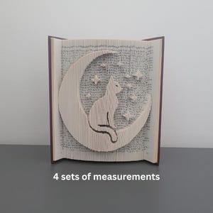 May include: A book with pages folded to create a cat silhouette sitting on a crescent moon with stars. The book has a brown spine and is displayed upright. The cat and moon are white against the book's pages.