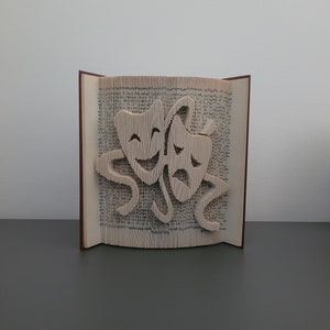 Könnte beinhalten: Ein Buch, dessen Seiten zu zwei Theatermasken gefaltet sind, eine lächelnd und eine grimmig. Die Masken sind aus Papier und mit Klebstoff am Buch befestigt.