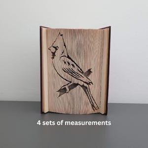 Peut inclure: Un livre décoratif avec un motif d'oiseau sculpté. L'oiseau est perché sur une branche, et les détails sont gravés dans les pages. Le livre a une couverture marron foncé et le texte "4 sets of measurements" est visible.