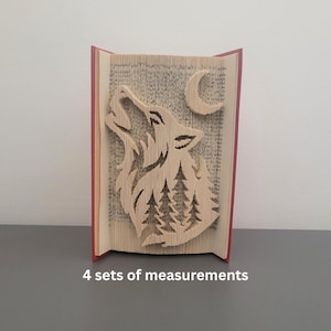 May include: A book sculpture featuring a howling wolf design. The wolf, trees, and crescent moon are carved from the pages of a book. The book has red edges and the text "4 sets of measurements" is at the bottom.