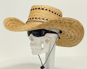 Può includere: Un cappello da cowboy in paglia intrecciata color marrone chiaro con tesa larga e curva, dotato di un cordino grigio sottogola con fermo nero. Il cappello è posizionato su un modello di teschio bianco che indossa occhiali da sole neri.