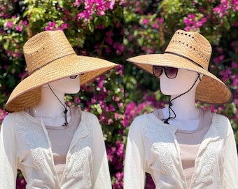 Pode incluir: Dois chapéus de palha de aba larga com uma correia para o queixo. Os chapéus são marrons e têm uma textura tecida. Os chapéus são exibidos em manequins contra um fundo de flores cor-de-rosa.
