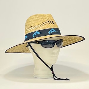 May include: A straw hat with a wide brim and a black band featuring a blue marlin design. The hat has a black chin strap and is worn on a mannequin head.