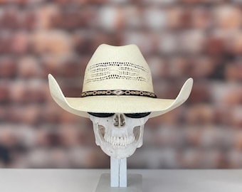 Può includere: Un cappello da cowboy in paglia color crema con corona ventilata e una fascia marrone scuro caratterizzata da un accento metallico centrale a forma di diamante. Il cappello è esposto su un supporto a forma di teschio bianco con occhiali.