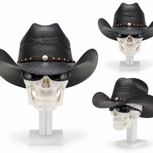 Op de afbeelding: Een zwarte cowboyhoed met een textuurafwerking, voorzien van een bruine leren band versierd met zilveren studs en een centrale zilveren concho. De hoed wordt getoond op een wit schedelmodel met een zwarte zonnebril op een witte voet.
