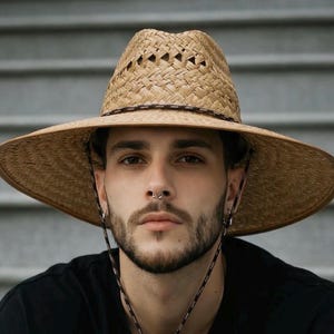 May include: A straw hat with a wide brim and a brown band. The hat has a woven design and a chin strap. The person is wearing a black shirt.