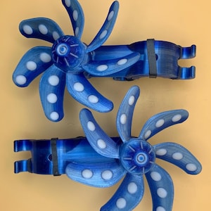 Puede incluir: Dos objetos decorativos azules y blancos en forma de abanico. Cada abanico tiene ocho aspas curvas con lunares blancos. Los abanicos están conectados a una base azul con una banda negra. Los objetos están sobre un fondo amarillo claro.