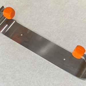 May include: A stainless steel, curved metal object with two orange, geometric knobs. The metal piece has a rectangular shape with curved ends and two small holes. The knobs are a bright orange color.