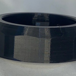 Op de afbeelding: Een zwarte, geometrische ring met een facetontwerp. De ring heeft een brede band en een gladde binnenkant. De ring is gemaakt van een solide, donker materiaal en lijkt 3D-geprint te zijn. De ring is een unisex accessoire.