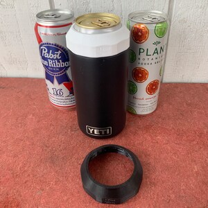 Op de afbeelding: Een zwarte YETI-blikjeskoeler met een witte bovenkant, naast drie drankblikjes. E&eacute;n blikje is een Pabst Blue Ribbon-bier en de andere is een Plant Botanical Vodka Seltzer. Een zwarte plastic ring ligt ervoor.
